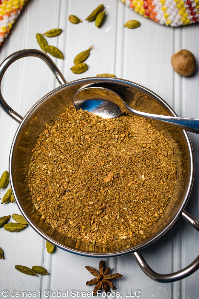 A silver bowl with handles filed with ground garam masala spice mix. Green cardamom pods, whole nutmegs, and a single whole star anise lay scattered on the grey wooden table.  A colorful orange, yellow, and white towel peaks in the top edge of the photo.